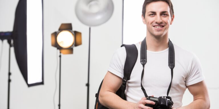 Man smiling, with his camera strap around his neck and studio lighting behind him.